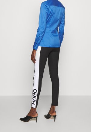Femme debout, portant des leggings noirs et blancs à rayures latérales avec le logo Hugo, un haut à manches longues bleu et des mules noires à talons.