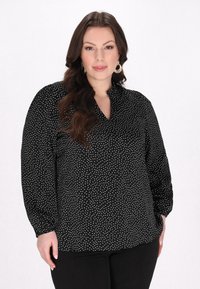 Schwarze Bluse mit weißen Punkten, mit Kragen und langen Ärmeln. Der Stoff ist glatt, hat eine lockere Passform und geraffte Bündchen.