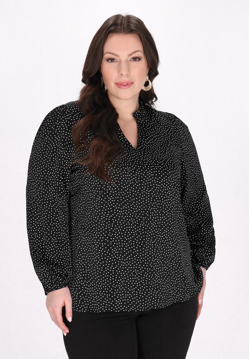 Schwarze Bluse mit weißen Punkten, mit Kragen und langen Ärmeln. Der Stoff ist glatt, hat eine lockere Passform und geraffte Bündchen.
