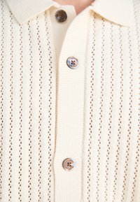 Knit polo shirt in light beige with a textured, open weave pattern. Features a pointed collar and two round, brown buttons.