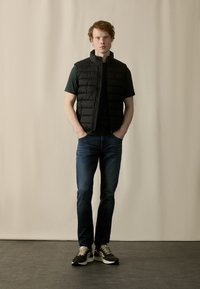 Sort dunvest over en grøn t-shirt, parret med blå jeans og brune sneakers. Sat mod en neutral baggrund. Glat tekstur.