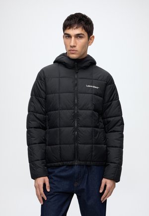 Jeune homme portant une veste matelassée noire à capuche avec le texte "Leave blank" sur la poitrine et un jean bleu, se tenant devant un fond clair.
