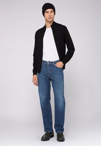 Svart zip-up jacka över en vit t-shirt, blå jeans och svarta skor. En enkel design med en figursydd silhuett och avslappnad passform i denim.