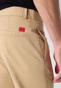 Pantalon beige avec une étiquette rouge "HUGO" au-dessus de la poche arrière, porté par une personne avec une chemise noire et la main droite visible.