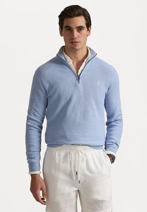 Light blue knitted quarter-zip sweater with ribbed cuffs and collar, worn over a white collared shirt, paired with white linen shorts.