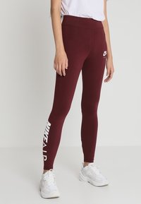 Bordeaux leggings gjorda av stretchmaterial, hög midja, med en vit Nike Air-logotyp längs vänster sida. Slät textur, figurnära design.