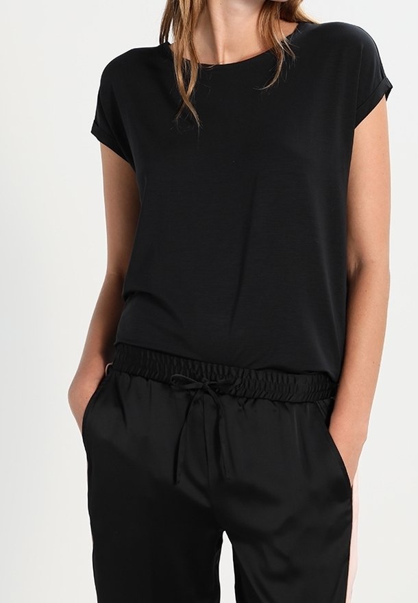 Kvinna iklädd en svart kortärmad topp och svarta byxor med dragsnöre, med händerna i fickorna mot en enfärgad bakgrund.