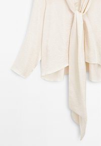Blusa color crema con una finitura testurizzata, caratterizzata da maniche lunghe e un dettaglio legato sul retro. L'orlo è asimmetrico con un design fluido.
