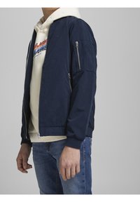 Man i en marinblå bomberjacka över en krämfärgad hoodie med grafik och blå jeans, stående mot en enkel grå bakgrund.