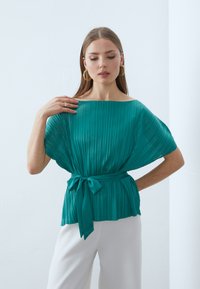 Blusa plissettata color turchese con scollatura a barca ampia e maniche corte. Presenta una cintura annodata in vita per un silhouette aderente. Tessuto morbido.
