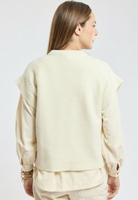 Femme aux cheveux bruns raides portant un pull sans manches en tricot crème par-dessus une chemise manches longues beige, vue de dos.