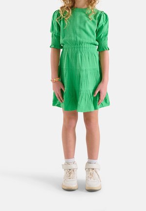 Robe verte à manches courtes avec tissu texturé, manches bouffantes smockées, taille froncée et jupe à volants. Portée avec des baskets blanches montantes.