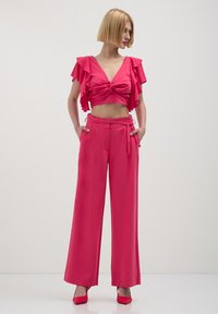 Femme aux cheveux blonds courts portant un crop top à volants rose à manches courtes et un pantalon large à taille haute assorti avec des talons roses.