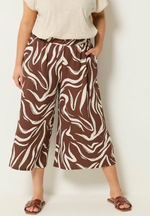 Vrouw draagt brede bruine broek met witte abstracte spiralen, beige top en bruine instapsandalen, staat met één hand in de zak.