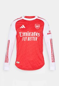 Crvena dugorukava majica s uzorkom, bijele rukave i crvene pruge. Sadrži Arsenalov logo i natpis "Emirates Fly Better".