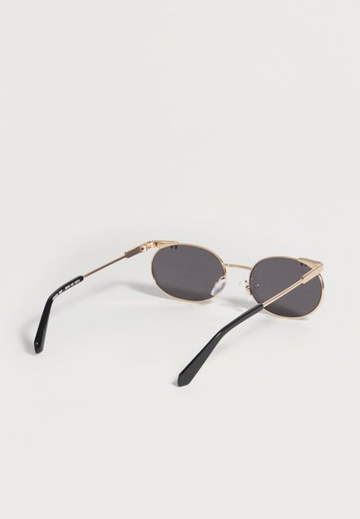 Lunettes de soleil ovales à monture dorée avec verres foncés et branches noires courbées, posées ouvertes sur une surface blanche.