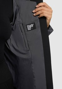 Chaqueta gris oscuro con un interior ligero y brillante. Cuenta con una etiqueta de marca negra con texto blanco. El diseño incluye una silueta ajustada y moderna.