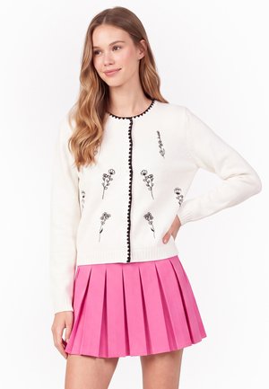 Donna che indossa un cardigan bianco con ricamo floreale nero e bordo smerlato abbinato a una gonna plissettata rosa acceso.