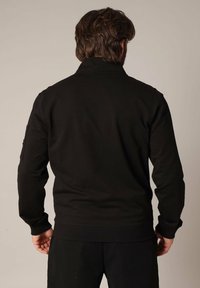 Zwarte trui met hoge kraag, lange mouwen en geribbelde manchetten. Voorzien van een zijzak op de linkermouw en een effen achterkant.