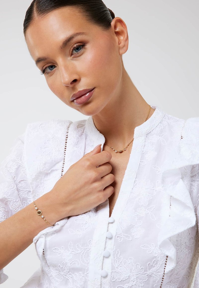 Witte geborduurde blouse met ruches aan de mouwen, knoopsluiting aan de voorkant en bloemmotief. Accessoires zoals een gouden armband en ketting voegen extra detail toe.