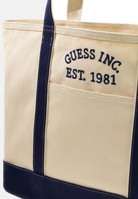 Borsa tote in tela beige con dettagli blu navy, caratterizzata da ricamo "GUESS INC. EST. 1981" e una tasca frontale per riporre oggetti.