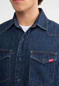 Camisa de mezclilla azul oscuro con un cuello clásico, dos bolsillos en el pecho, costuras visibles y una pequeña etiqueta de marca roja en el bolsillo izquierdo.