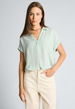 Jeune femme aux longs cheveux bruns portant un chemisier vert clair à manches courtes et un pantalon taille haute couleur crème, une main dans la poche.