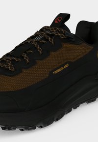 Gros plan d'une basket Timberland noire et marron avec des lacets à motifs et une semelle épaisse à crampons sur fond blanc.