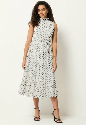 Femme aux cheveux bouclés portant une robe midi blanche sans manches, à pois noirs, avec une ceinture à nouer, assortie à des sandales noires à talons hauts.