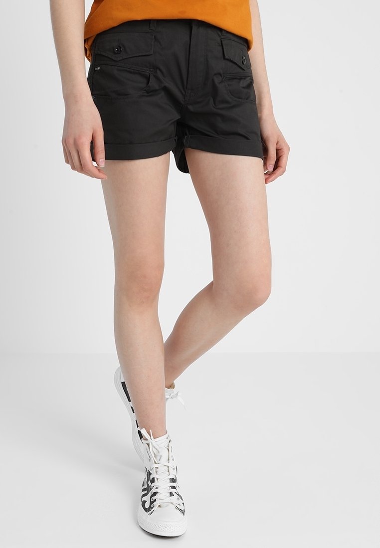 Personne portant un short noir avec revers et poches à boutons, un haut orange, et des baskets montantes blanches avec des motifs de flammes noires.
