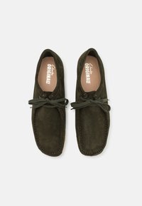 Tummanvihreät mokkanahkaiset mokkasiinijalkineet, joissa on nauhakiinnitys, korostettuna ommelluilla yksityiskohdilla ja vaaleanruskea sisusta. Litteä pohja rentoon käyttöön.