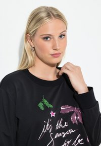 Sudadera negra con cuello redondo, que presenta un estampado multicolor de texto y diseños, incluyendo lentejuelas y un detalle de cinta.