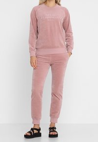 Chándal de dos piezas de terciopelo rosa que presenta un sudadero con cuello redondo y logotipo en relieve, acompañado de pantalones de chándal ajustados a juego. Superficie plana y texturizada.