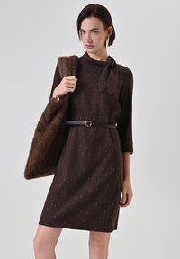 Robe marron avec un motif subtil, manches longues et un détail de col noué ; accessoirisée d'un châle marron moelleux et d'une fine ceinture.