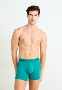 Zöldeszöld pamut boxeralsó, fekete Calvin Klein derékpánttal, testhezálló fazonnal és sima tapintással. A modell egy sima háttér előtt áll.
