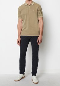Olivově zelené polo tričko s límečkem, krátkým rukávem a malým logem; v kombinaci s námořnickými slim-fit kalhotami a bílými teniskami s hnědými akcenty.