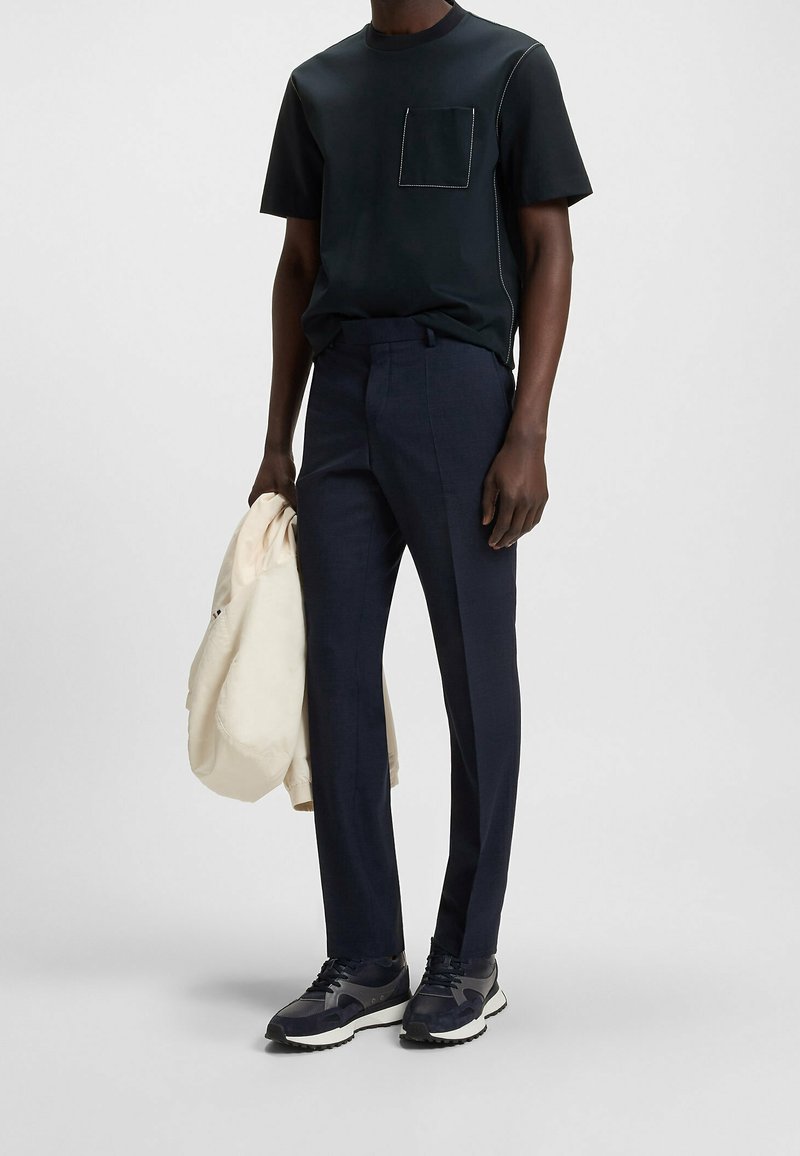 Homme portant un t-shirt noir avec une poche, un pantalon slim bleu marine, des baskets foncées, tenant une veste beige clair dans une main, debout sur un fond uni.