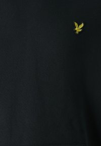 Sudadera negra con un pequeño logotipo de águila bordado en amarillo en la parte superior izquierda. La tela parece suave y tiene un corte clásico.