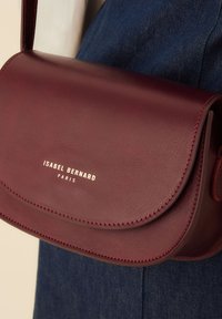 Burgunderfarbene Ledertasche mit Umhängeriemen, Klappenverschluss, goldgeprägtem Logo und sichtbaren Steppnähten. Glatte Textur und kompakte Form.