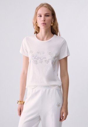 Donna bionda che indossa una camicia bianca a maniche corte con motivo floreale argento e pantaloni bianchi, accessoriata con grandi orecchini e braccialetti dorati.