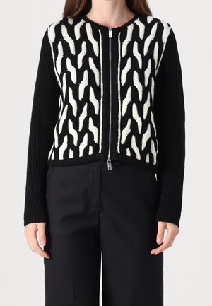 Frau trägt eine schwarze Strickjacke mit Reißverschluss, vorne mit weißen, strukturierten Wellenmustern und an Ärmeln sowie Saum in einfarbigem, schwarzem Rippstrick.