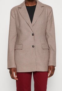 Blazer con un patrón a cuadros marrón y crema, cierre de un solo botón, solapas con muesca y dos bolsillos laterales. Llevado sobre una blusa negra.