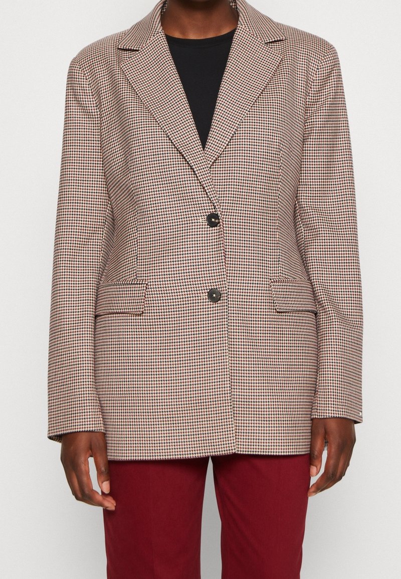 Blazer con un patrón a cuadros marrón y crema, cierre de un solo botón, solapas con muesca y dos bolsillos laterales. Llevado sobre una blusa negra.