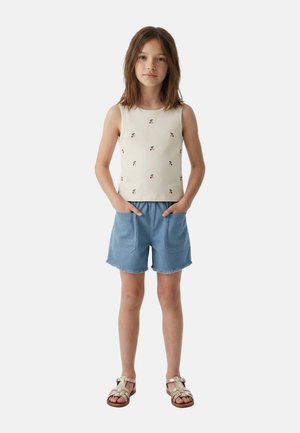 Fille debout, mains dans les poches, portant un débardeur beige avec imprimé cerises, un short bleu avec poches avant et des sandales beiges.