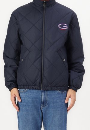 Person iført en marineblå quiltet jakke med frontlynlås, elastiske manchetter og logoen "G" på brystet, der er parret med blå jeans.