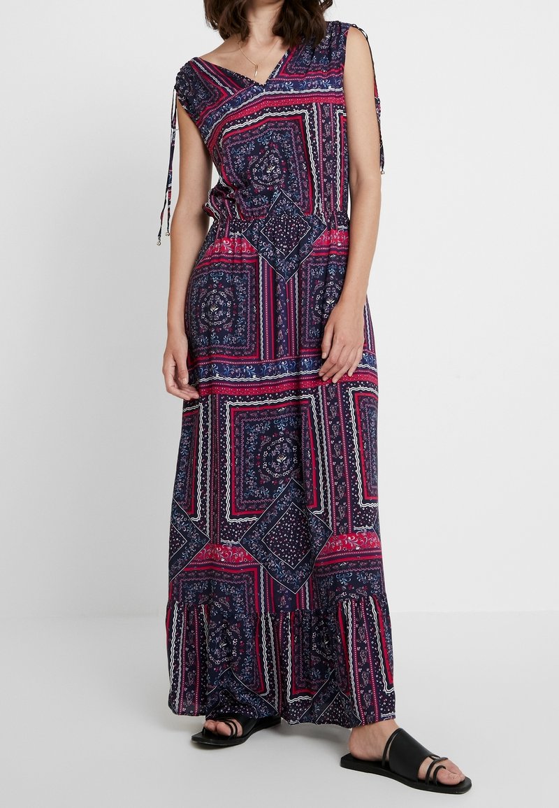 Femme portant une robe maxi sans manches avec des motifs géométriques et floraux bleu marine, rose et blanc, associée à des sandales noires ouvertes à l'avant.