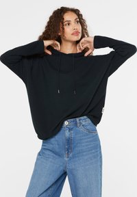 Schwarzer Kapuzenpullover mit gerippter Textur, lockerer Passform und Zugbanddetails, kombiniert mit hellblauen hochtaillierten Jeans.