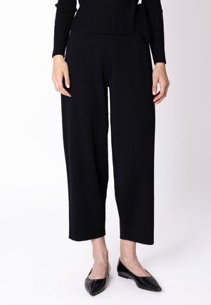 Donna che indossa pantaloni neri a gamba larga e corta, ballerine nere con punta affilata e un top nero a maniche lunghe a coste con cintura in vita.