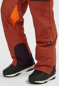 Pantalones resistentes al agua de color rojo ladrillo con acentos negros. Cuentan con un bolsillo de malla y cierres a presión en el tobillo. El material es suave y duradero.