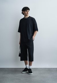 Uomo in piedi contro un muro grigio, indossa una t-shirt nera oversize, pantaloncini neri lunghi, calzini bianchi, sneakers nere, tiene una piccola borsa nera.